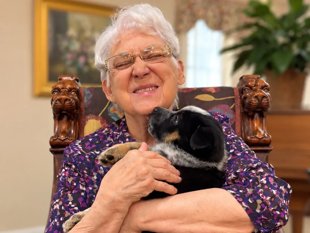 A smiling resident hugging a black puppy in her armchair