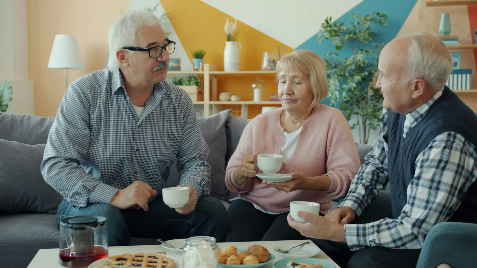 Three older friends laughing over coffee and treats on a sofa